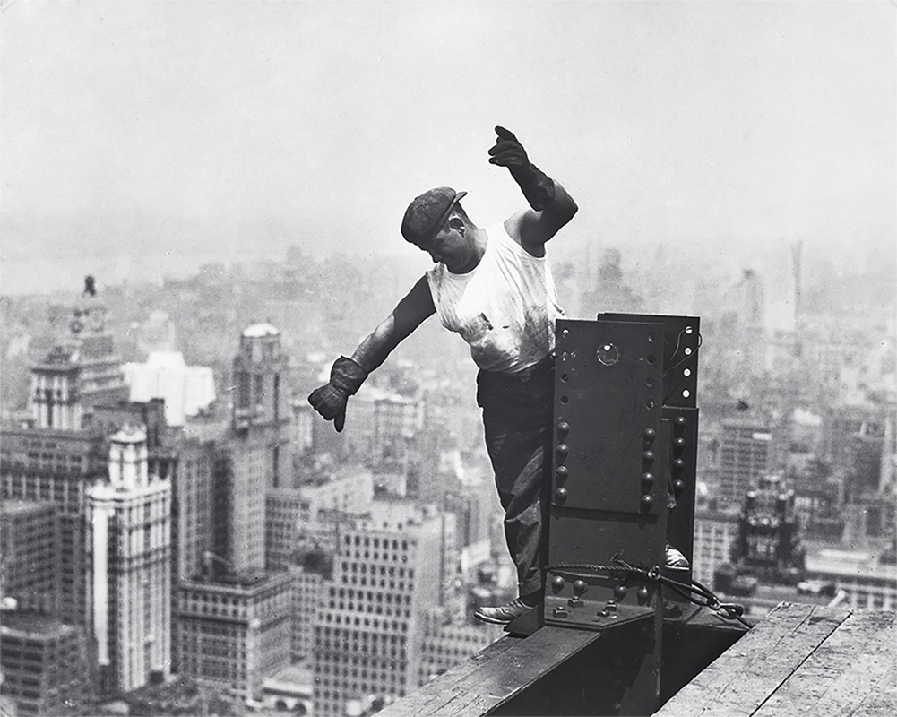 Black and white photograph of a construction worker in the sky, building the Empire State Building. View of New York skyscrapers behind.