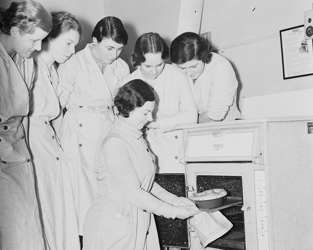 Black and white photograph of five women students stand watching as their domestic cooking class teacher kneels taking a pie baking out of the oven.
