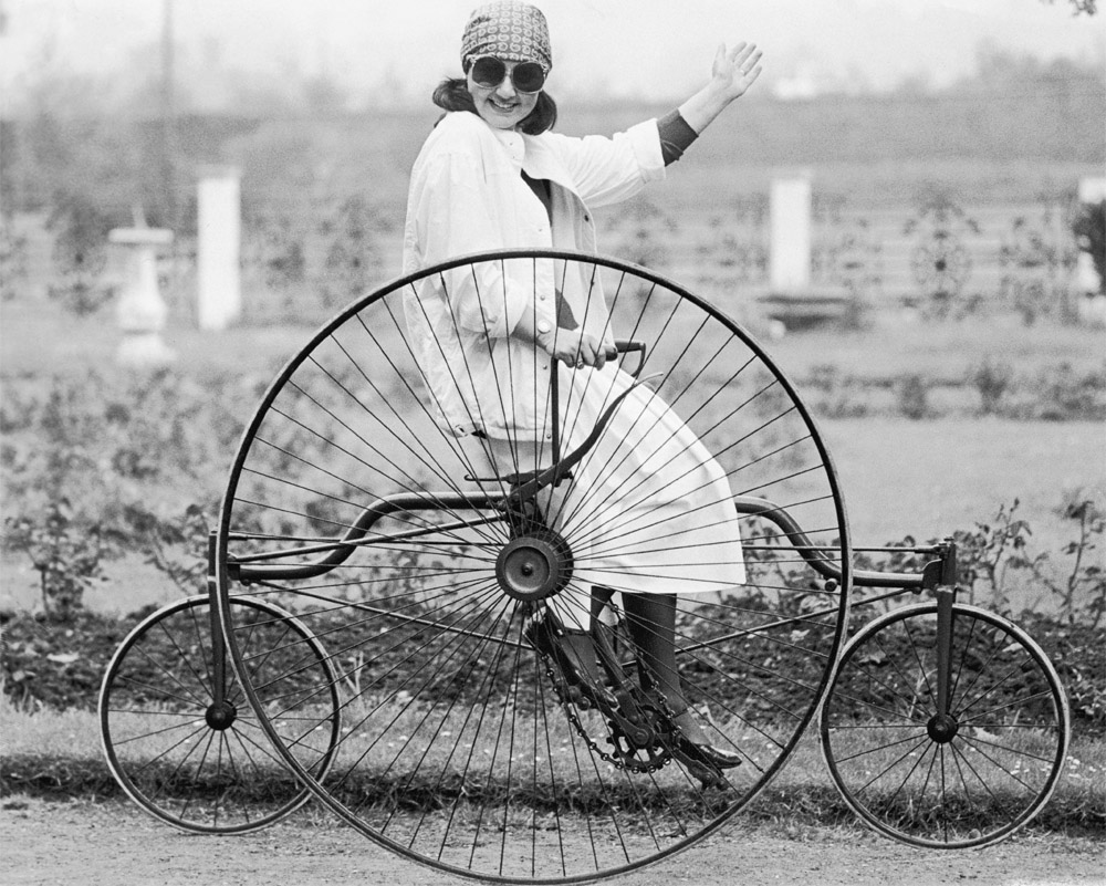 Black and white photograph of a woman on a tricycle, smiling, waving, wearing a white outfit and sunglasses
